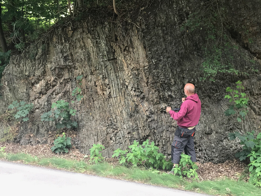 Der Fachschullehrer erläutert an einem Gesteinsaufschluss die geologische Entwicklungsgeschichte in diesem Gebiet. Entlang einer Straße steht Gestein mit einer senkrechten Böschung an.