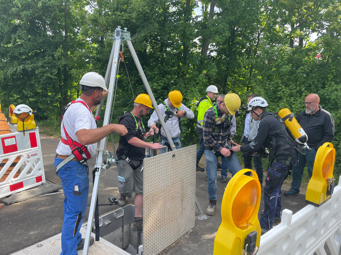 Eine Gruppe von drei Fachschulstudenten bereitet sich auf die Befahrung eines Brunnenschachtes vor. Zwei Fachschulstudenten haben bereits einen Höhenrettungsgurt angelegt. Ein Mitarbeiter de Sanierungsunternehmens ist dabei behilflich.